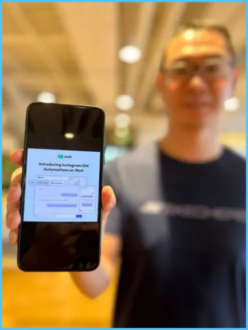 A person holding a phone toward the camera displaying a Wati screen about Instagram DM automation, with the person blurred in the background.